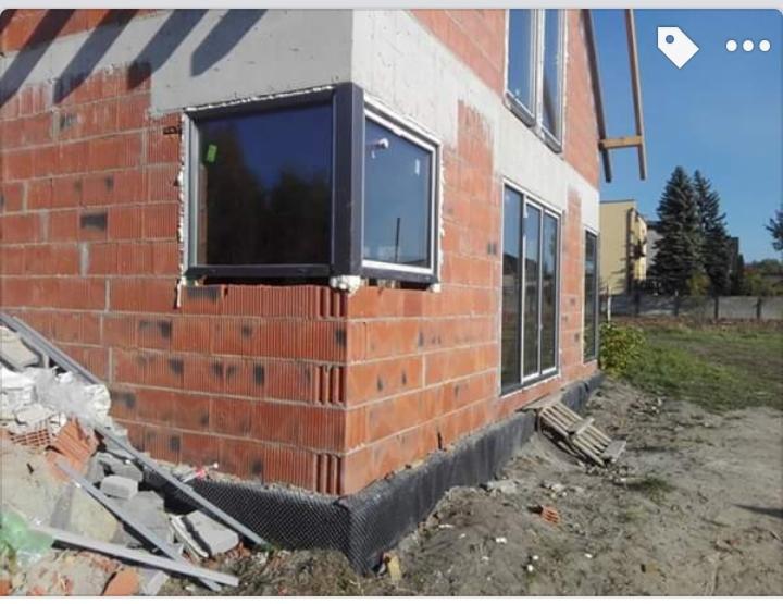 Widok z zewnątrz na budynek w trakcie montażu okien z czarnymi ramami, widoczny mur z czerwonej cegły, czarna izolacja fundamentu i częściowo zagospodarowany teren.