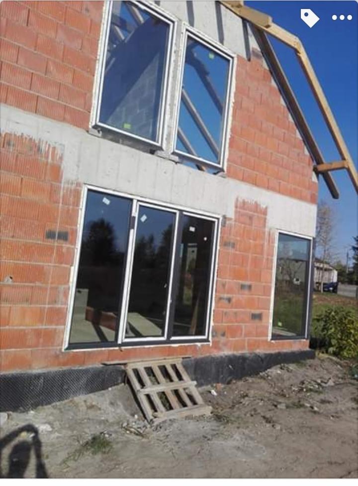 Surowy stan budynku z czerwonej cegły z zamontowanymi oknami PCV z czarnymi ramami; widoczne rusztowania i tymczasowe schodki z desek.