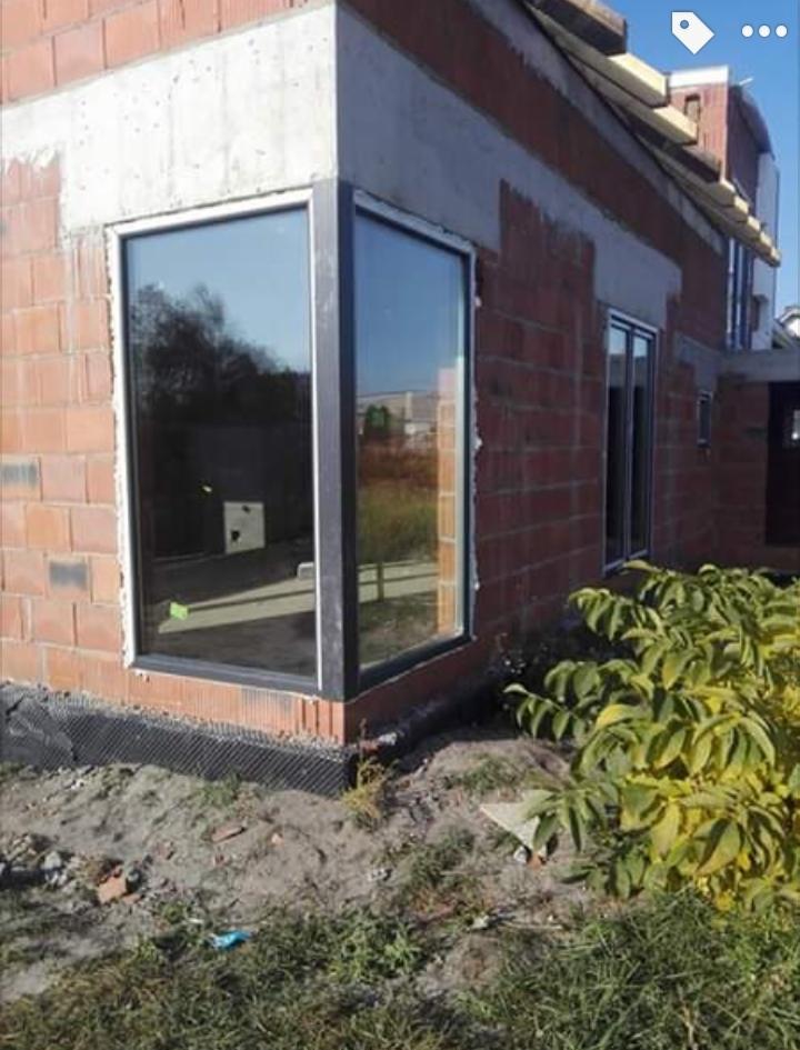 Nowe, duże okno narożne z czarną ramą w surowym stanie budynku z cegły, widoczne czarne zabezpieczenie fundamentów i ziemia.