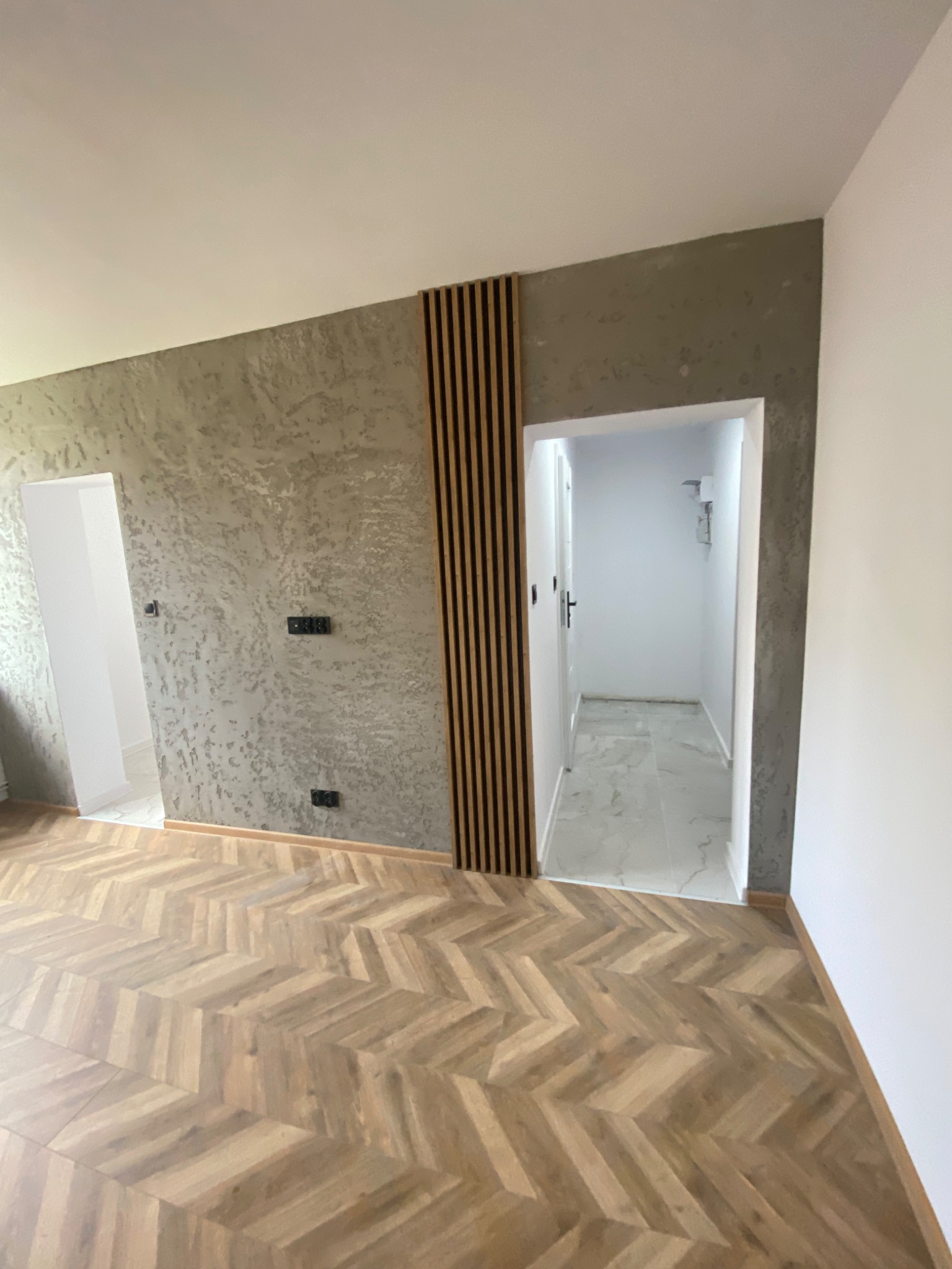 Wykończone wnętrze w Dydni: parkiet w jodełkę, dekoracyjna ściana z drewnianymi lamelami i fakturą betonu, widok na korytarz z marmurową podłogą.