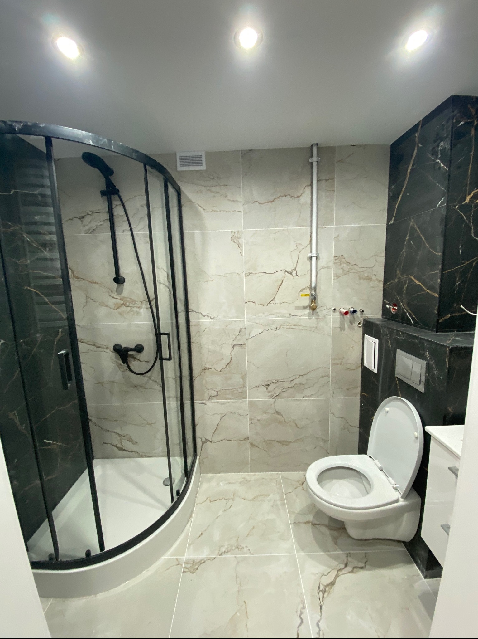 Wykończona łazienka z kabiną prysznicową typu walk-in z czarnymi profilami, toaletą podwieszaną i marmurowymi płytkami w beżach i czerniach.