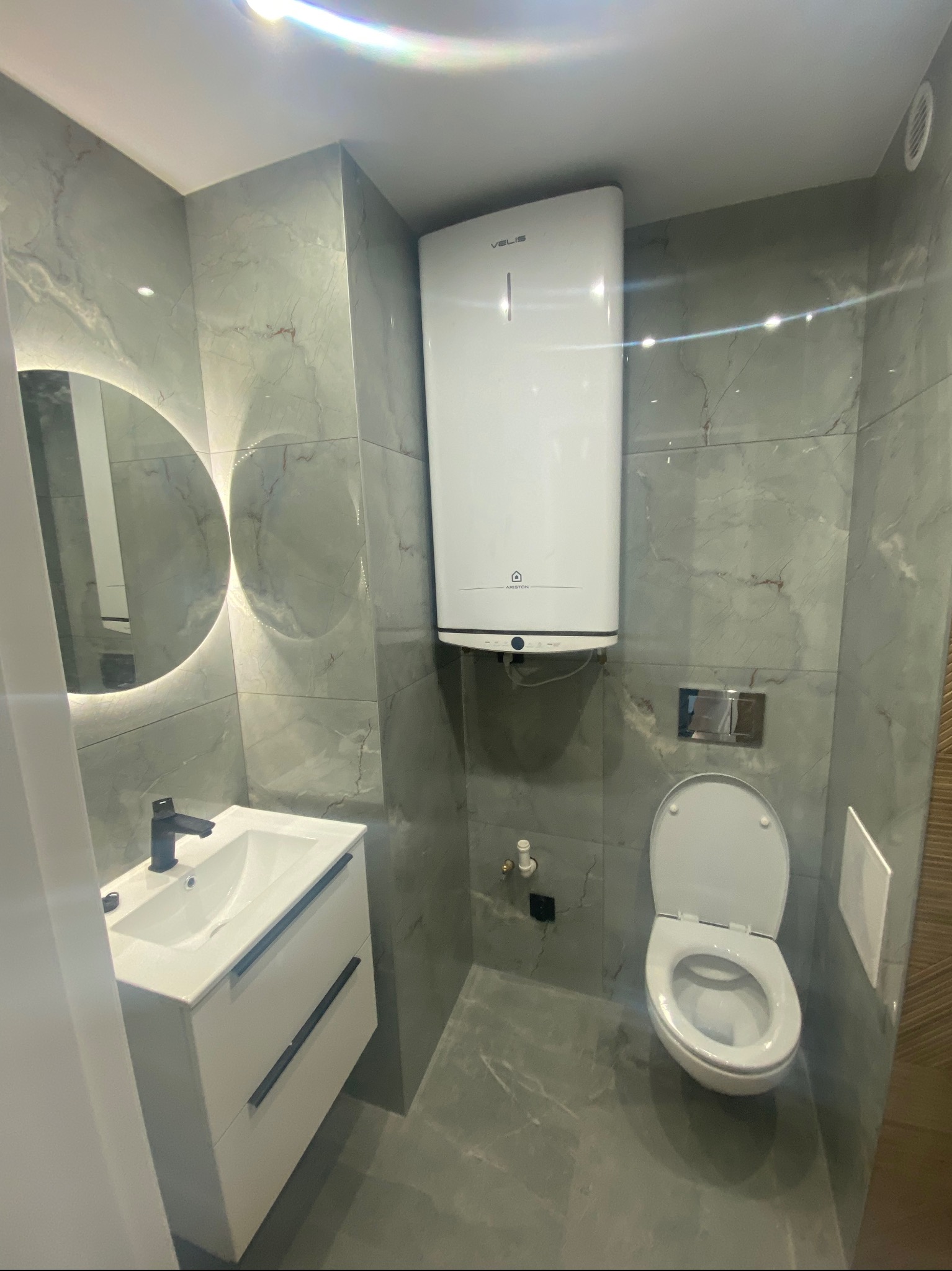 Nowoczesna łazienka z szarymi płytkami imitującymi marmur, podświetlanym lustrem i białymi meblami łazienkowymi. Widoczny bojler i toaleta.