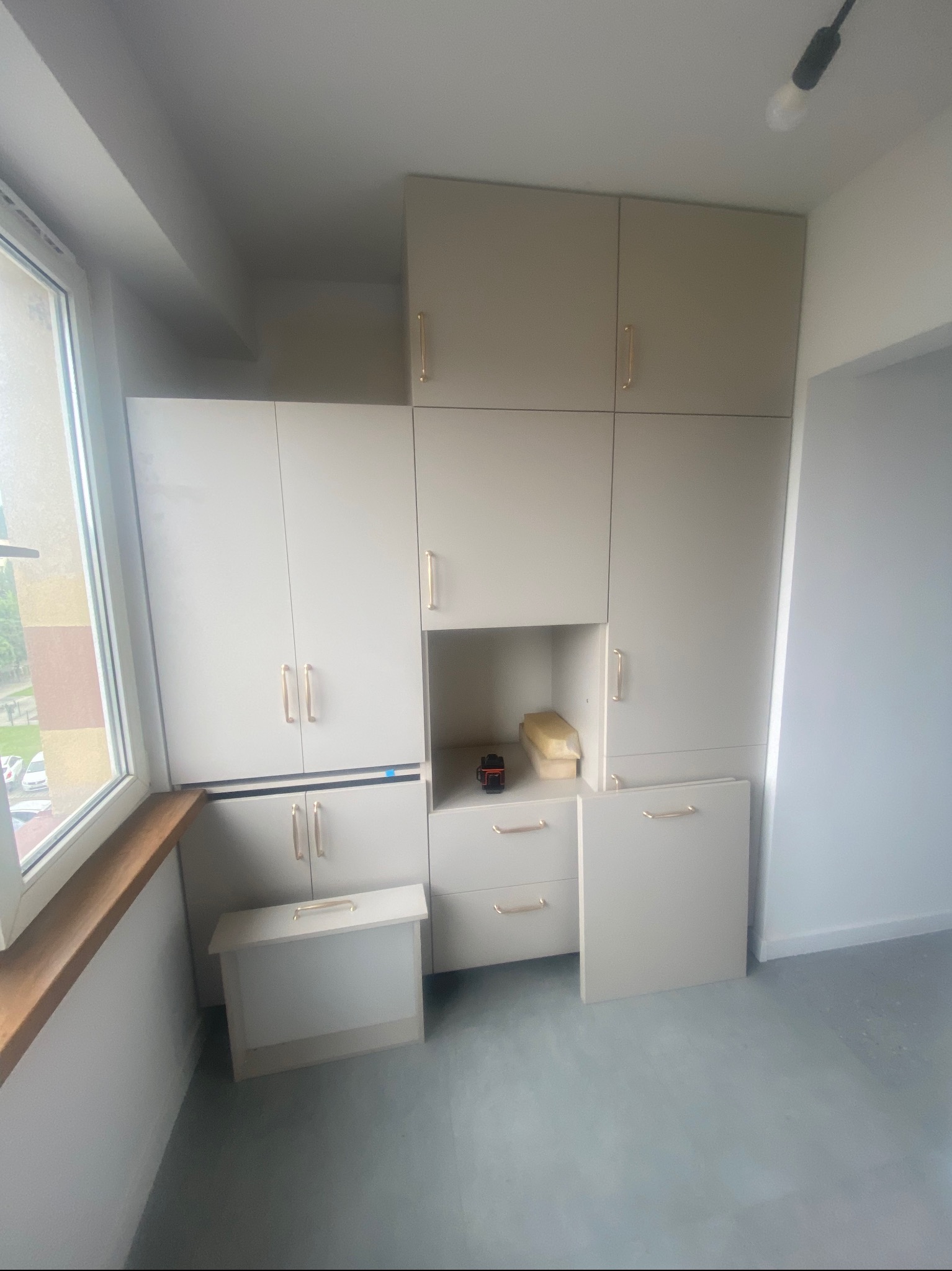 Jasne, modułowe szafy z złotymi uchwytami, zabudowane od podłogi do sufitu, z otwartą półką i szufladami, w pomieszczeniu z oknem i szarą podłogą.