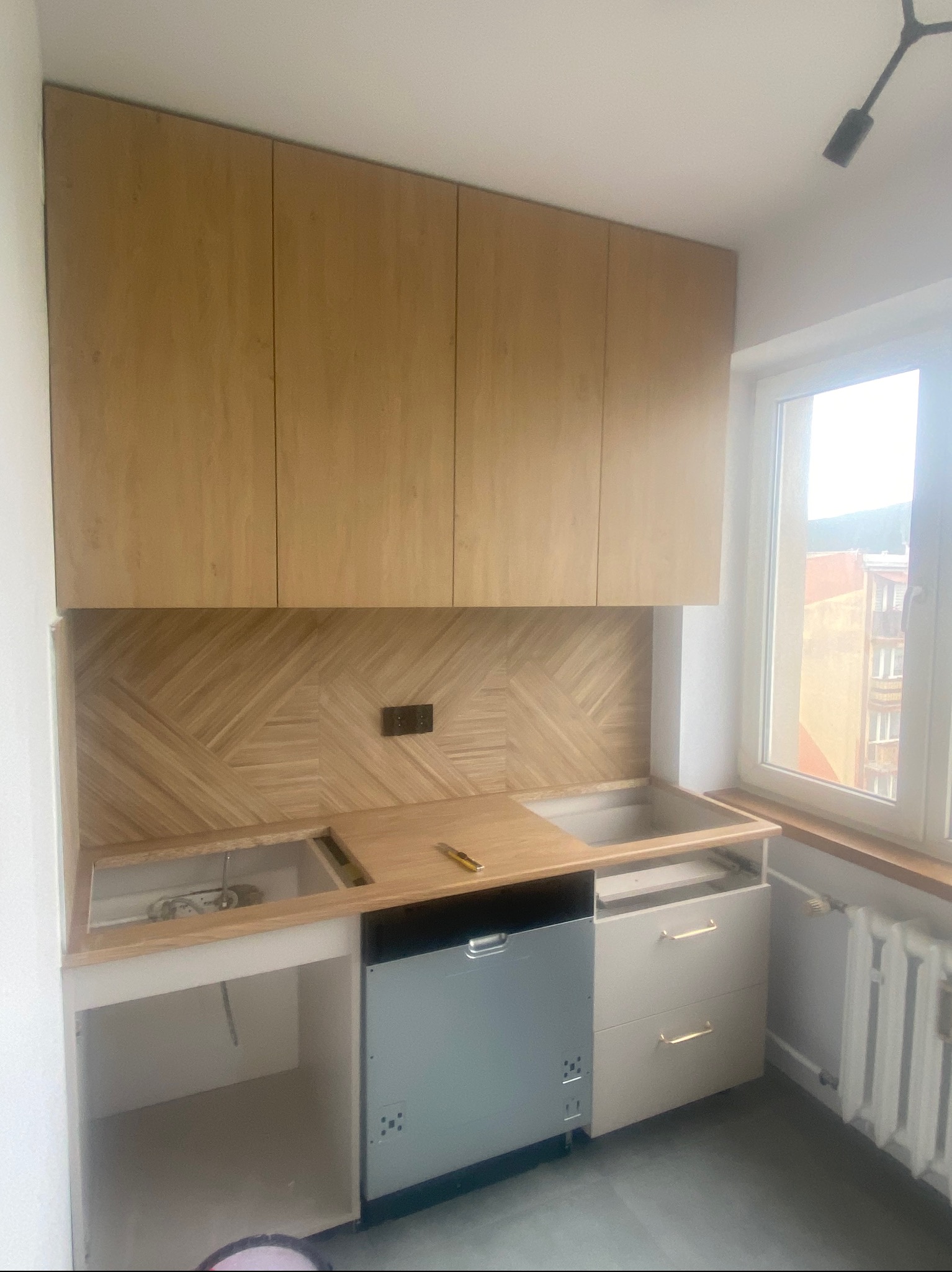 Jasna, drewniana zabudowa kuchenna z wzorem jodełki na ścianie, widoczna w trakcie montażu. Częściowo zamontowany sprzęt AGD i szuflady. Okno z widokiem na budynek.