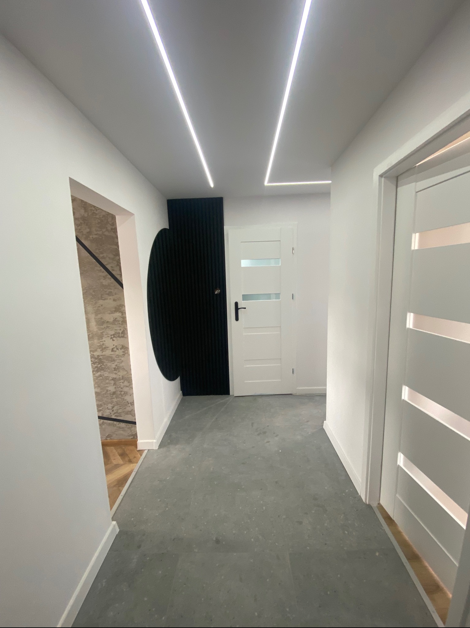 Nowoczesny korytarz z szarą posadzką, białymi ścianami, oświetleniem LED i dekoracyjnymi elementami: czarnym panelem oraz wykończeniem ściany z wzorem.