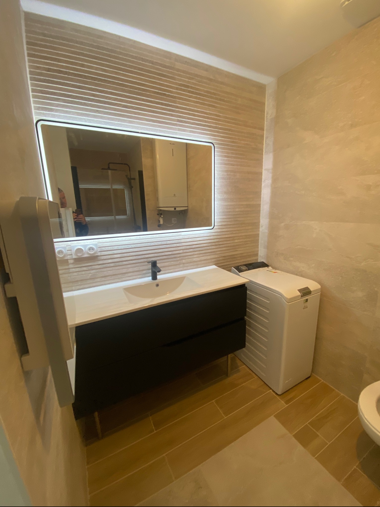 Nowoczesna łazienka w beżowych tonacjach z podświetlanym lustrem, umywalką z szafką i pralką. Widoczne płytki imitujące drewno na ścianach i podłodze.