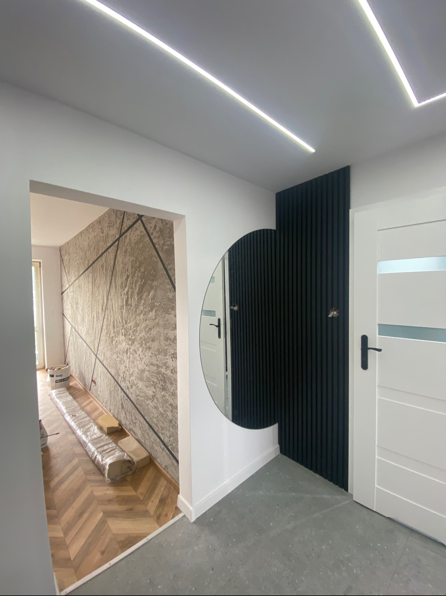 Nowoczesne wykończenie wnętrza: dekoracyjna ściana z geometrycznym wzorem, okrągłe lustro i czarne lamele obok białych drzwi. Oświetlenie LED na suficie.