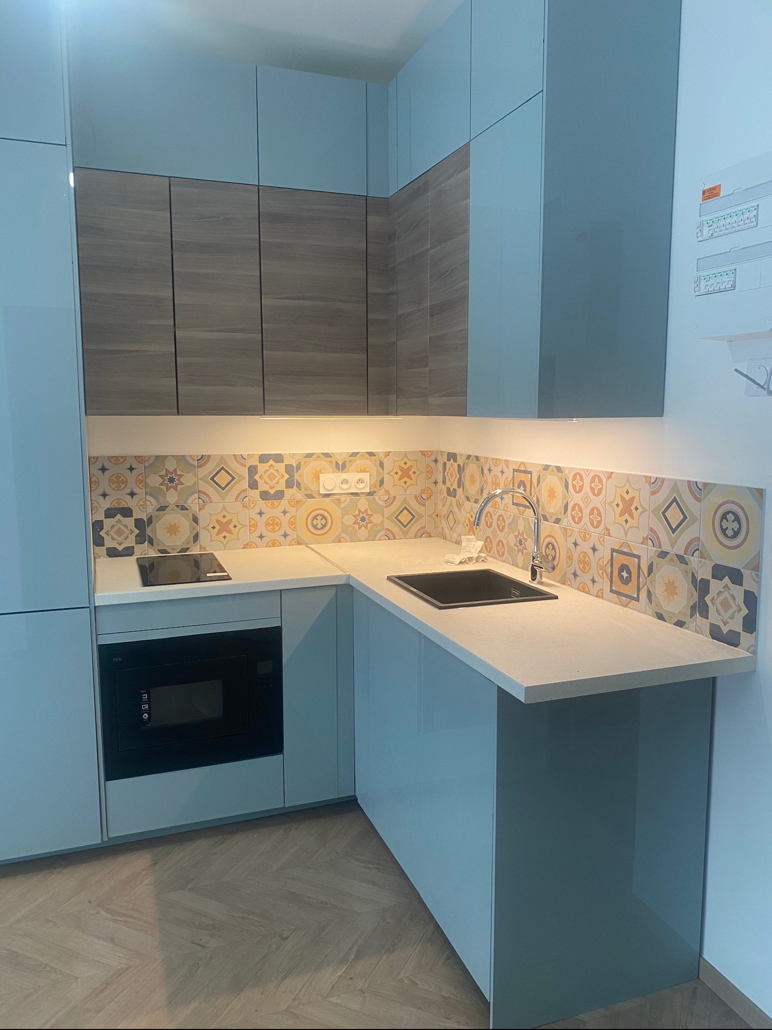 Niebieska zabudowa kuchenna z drewnianymi szafkami górnymi i wzorzystą mozaiką nad blatem. Widoczna płyta indukcyjna i piekarnik. Parkiet ułożony w jodełkę.