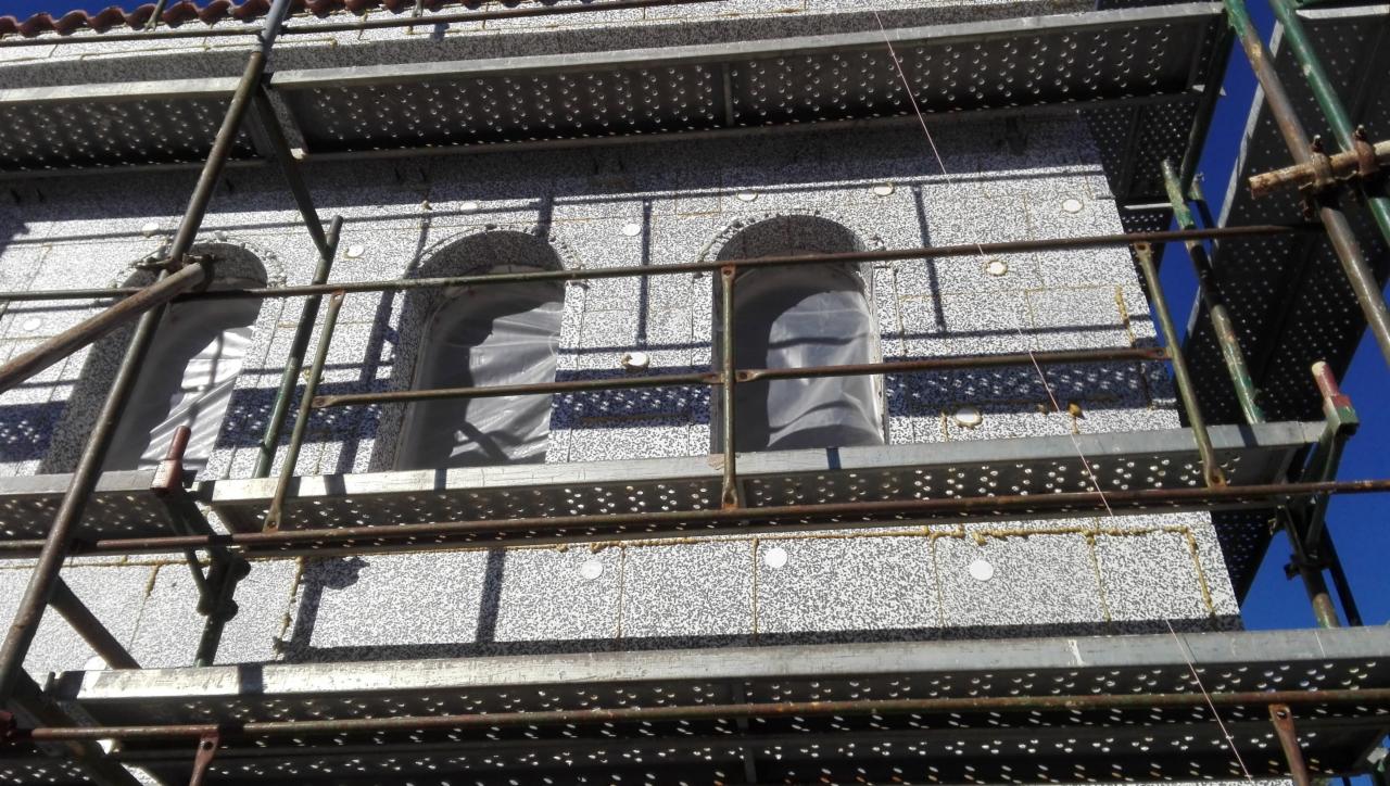 Budynek w trakcie ocieplania elewacji styropianem, widoczne rusztowania i zabezpieczone okna folią.