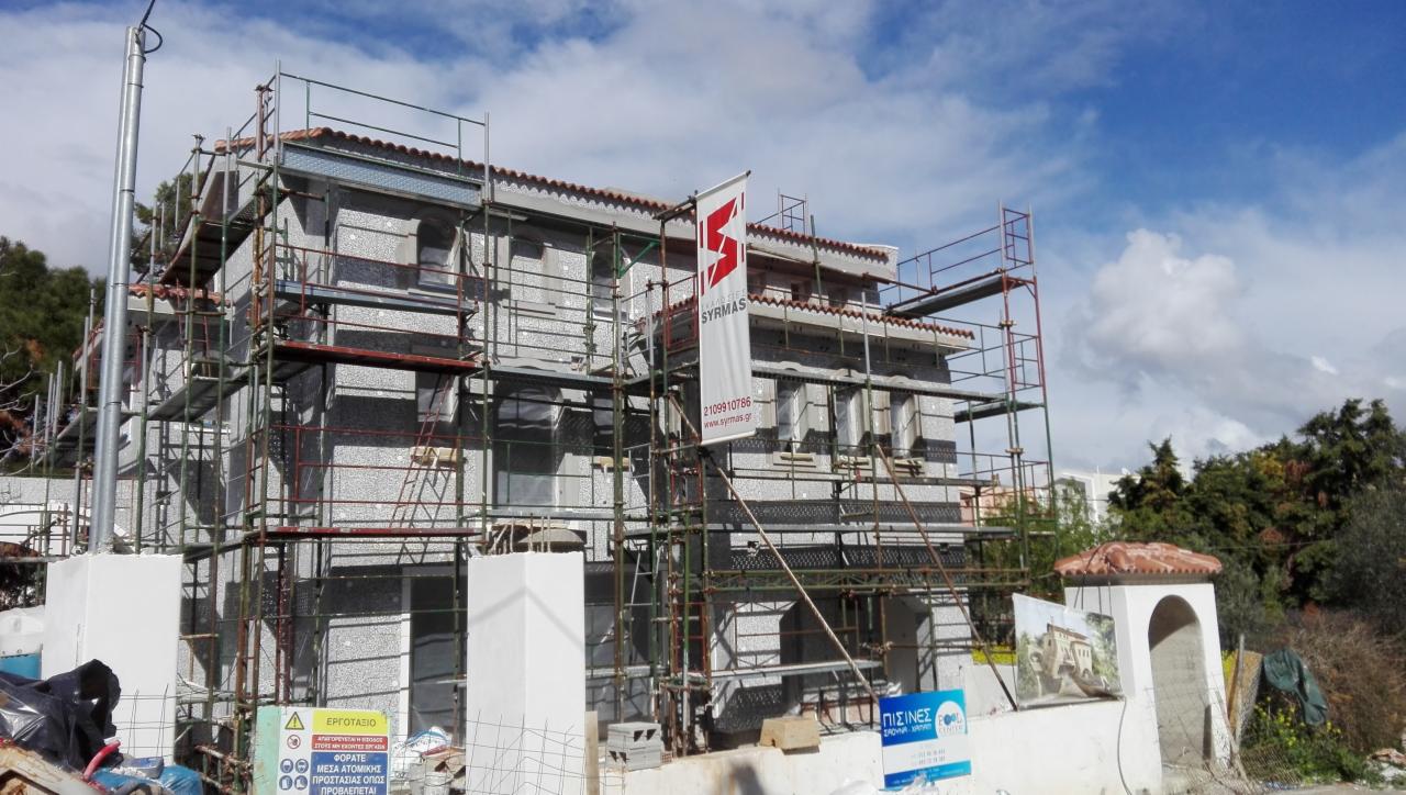 Budynek w trakcie ocieplania elewacji zewnętrznej, z widocznymi rusztowaniami, warstwą izolacyjną i banerem firmy na tle błękitnego nieba.