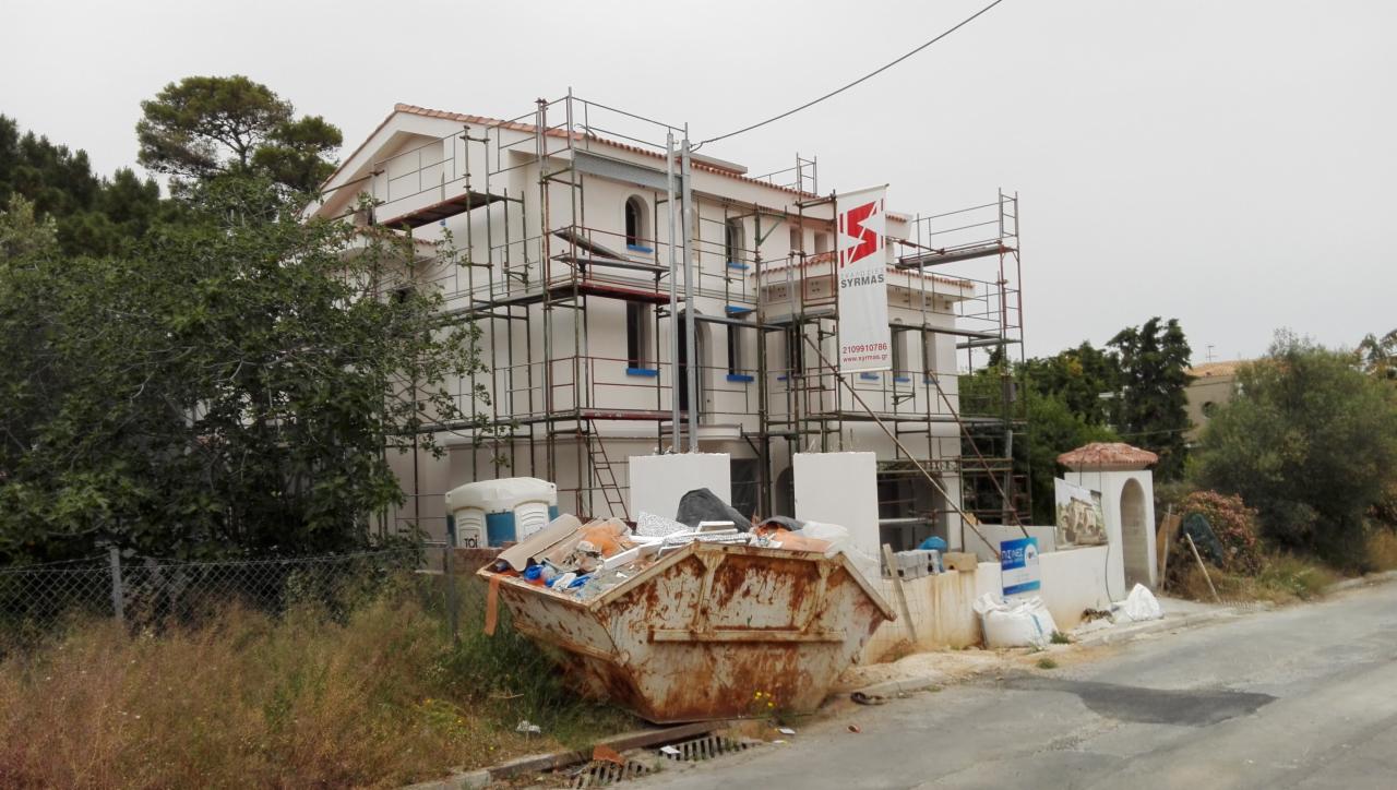 Dwupiętrowy dom w trakcie prac elewacyjnych, otoczony rusztowaniami, z widocznym kontenerem na odpady budowlane i banerem reklamowym firmy.