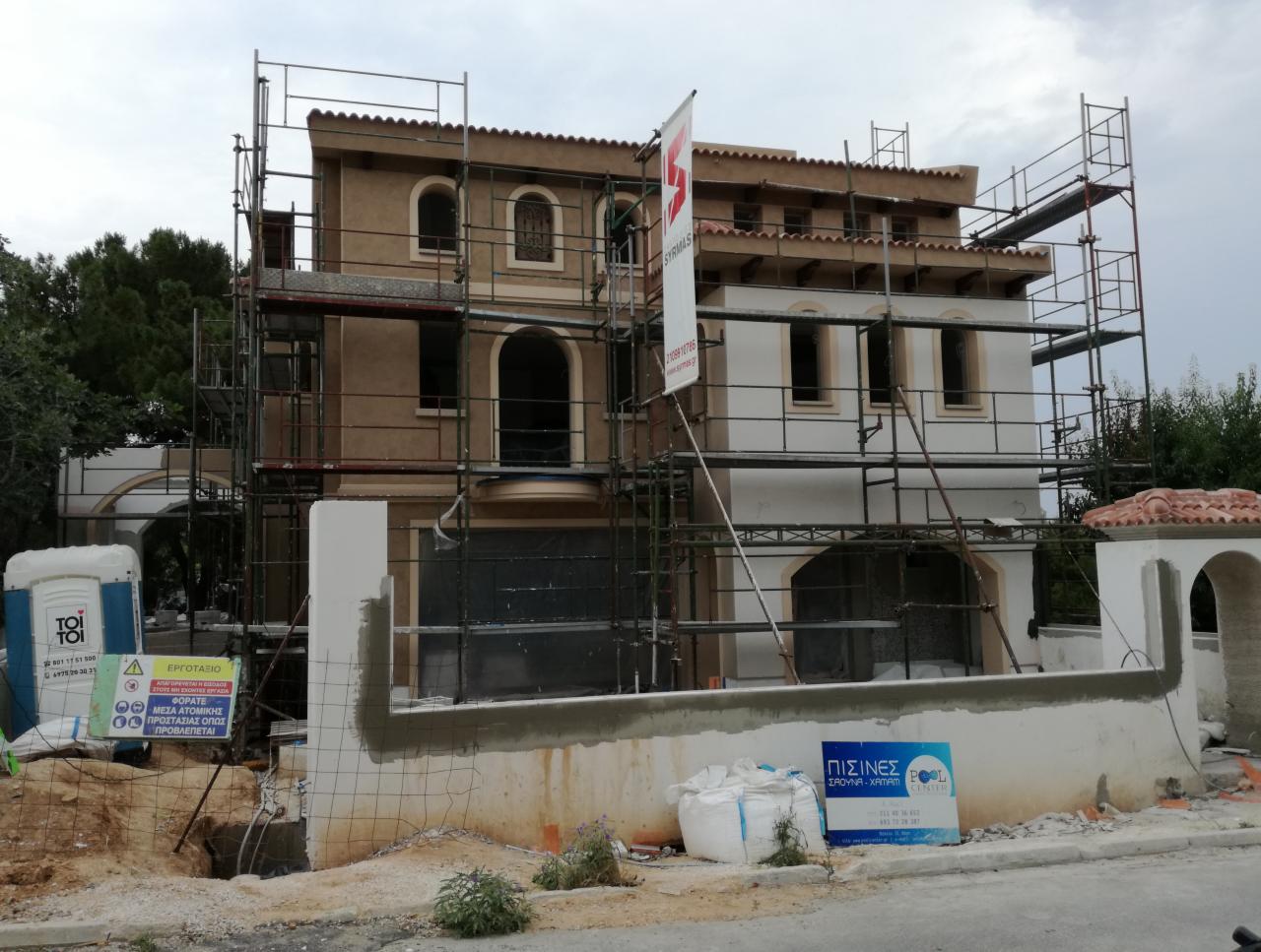 Dom w trakcie prac elewacyjnych, z widocznymi rusztowaniami, częściowo otynkowany, z reklamą firmy na ogrodzeniu i przenośną toaletą na placu budowy.