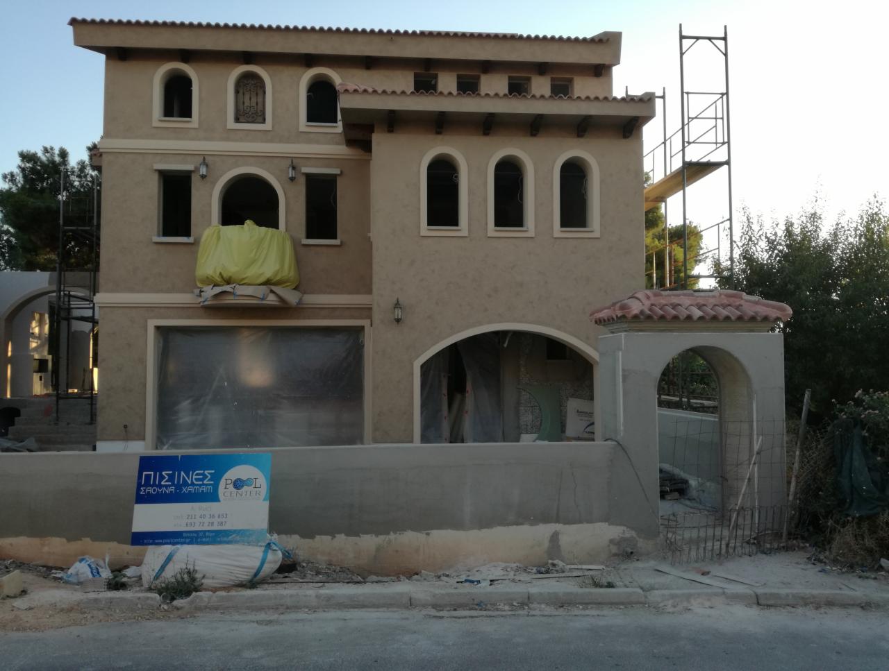 Dom w trakcie ocieplania elewacji, widoczne rusztowanie, okna zabezpieczone folią, na balkonie żółta plandeka, w tle zieleń i błękitne niebo.