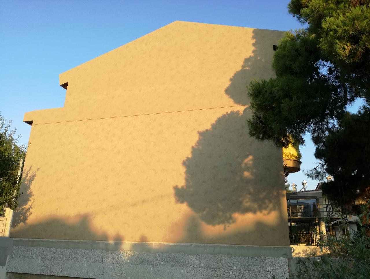 Żółta elewacja domu jednorodzinnego w trakcie prac, z widocznym ociepleniem fundamentów i cieniem drzewa na ścianie.