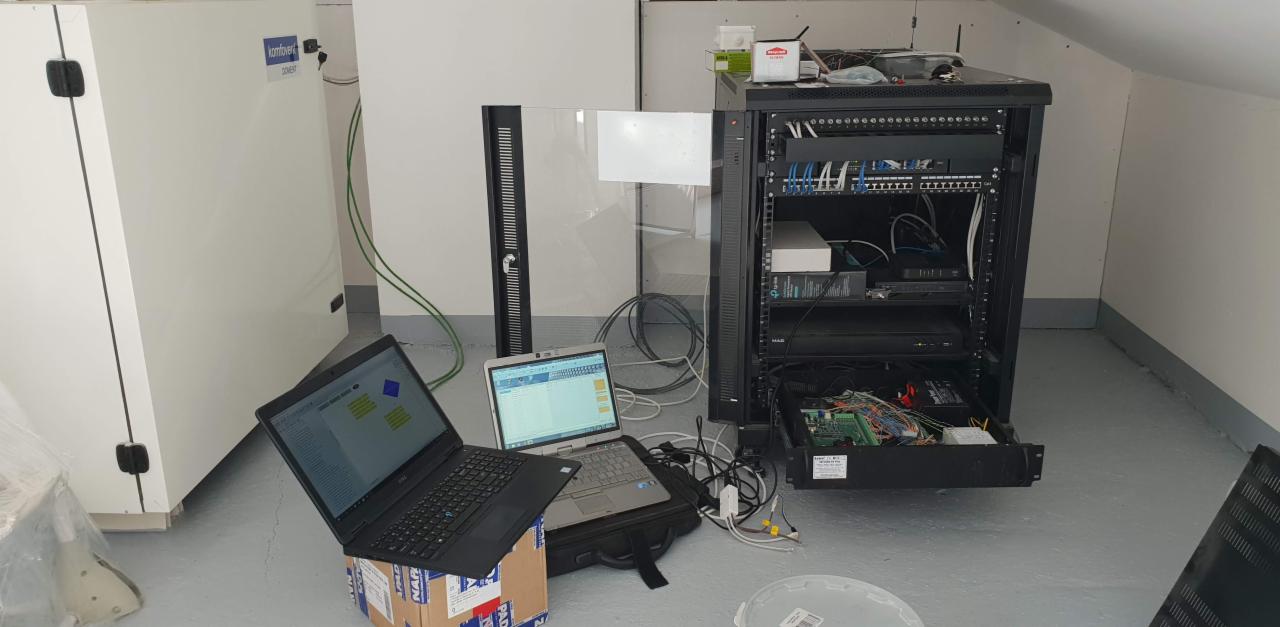 Serwerownia w trakcie konfiguracji: widoczna szafa rack z okablowaniem, dwa laptopy na kartonowym pudle i walizce obok, biała szafa telekomunikacyjna w tle.