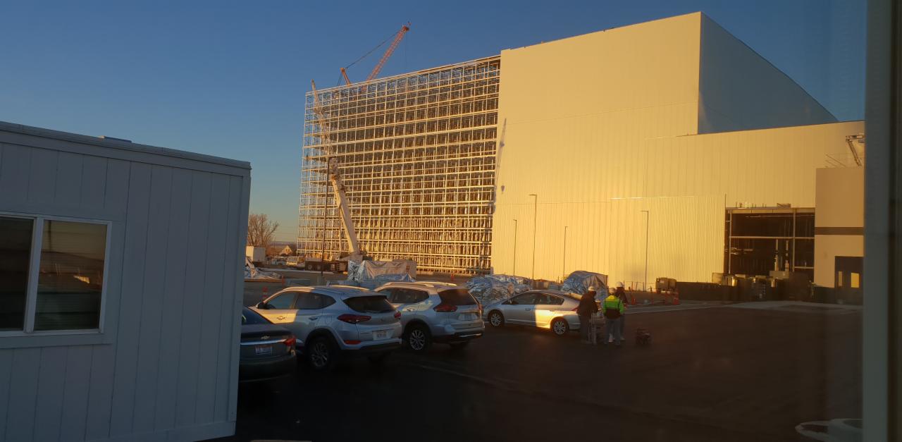 Budowa hali przemysłowej w trakcie montażu konstrukcji stalowej, widoczny żuraw, zaparkowane samochody i pracownicy w kamizelkach odblaskowych.