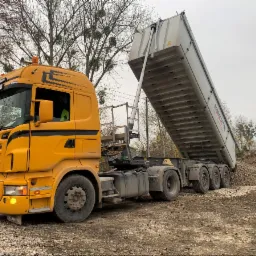 Żółta ciężarówka Scania z podniesioną naczepą wyładowuje materiał na ziemię, widoczna pryzma materiału.