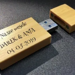 Drewniany pendrive z grawerem 'Nasze wesele DAREK & ANIA 04-05-2019', leżący na ciemnej, teksturowanej powierzchni z odłączoną zatyczką.