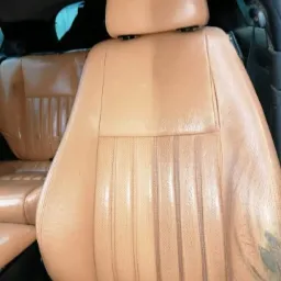 Fragment beżowego skórzanego fotela samochodowego, widoczna różnica w czystości po lewej stronie, z prawej strony widoczne ślady zużycia i zabrudzenia.