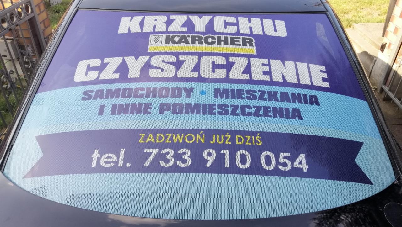 Reklama firmy sprzątającej Krzychu Karcher, naklejona na tylnej szybie samochodu, oferującej czyszczenie samochodów, mieszkań i innych pomieszczeń, z numerem telefonu.