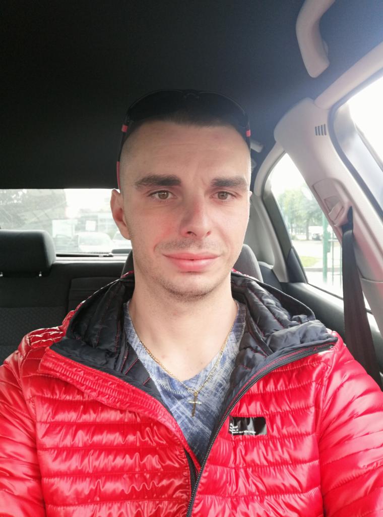 Mężczyzna w czerwonej kurtce, siedzący w samochodzie, patrzy w obiektyw. Widoczne wnętrze pojazdu i fragment ulicy w tle. Ujęcie z perspektywy pasażera.