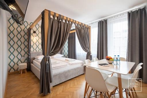 Sypialnia z łóżkiem z baldachimem, zasłony w odcieniach szarości, stół z owocami i wodą mineralną, parkiet w jodełkę, ściana z geometrycznym wzorem.