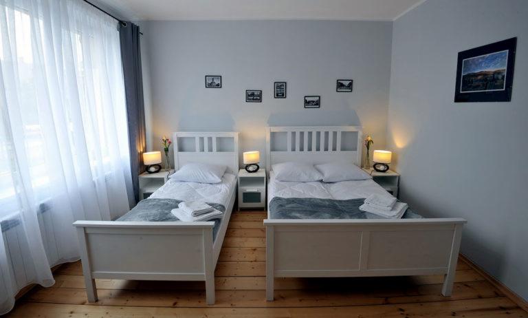 Pokój z dwoma białymi łóżkami, szafkami nocnymi z lampkami, zasłonami i drewnianą podłogą; minimalistyczny wystrój z obrazkami na ścianie.