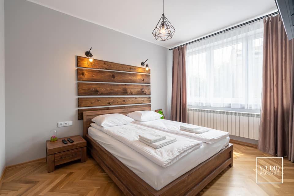 Nowoczesna sypialnia z drewnianym łóżkiem i zagłówkiem z desek podświetlonych kinkietami, brązowe zasłony przy oknie z białymi firankami, parkiet w jodełkę.