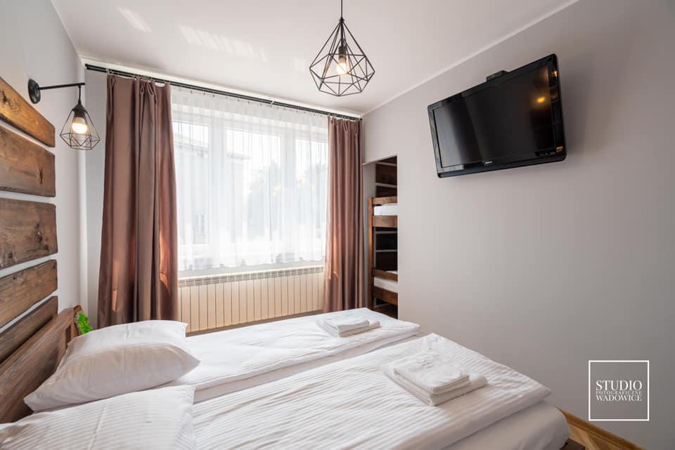 Jasne wnętrze sypialni z dwoma łóżkami, brązowymi zasłonami przy oknie z białymi firankami, geometryczną lampą wiszącą i telewizorem na ścianie.