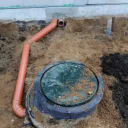 Montaż zbiornika z zieloną pokrywą i pomarańczową rurą odpływową, widoczne ślady ziemi na pokrywie i wokół zbiornika, świeży wykop przy fundamencie budynku z bloczków.