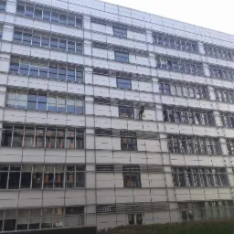 Pracownik na wysokości myje okna nowoczesnego biurowca z białymi panelami elewacyjnymi, widoczny z dołu, z żółtym wiadrem na linie.