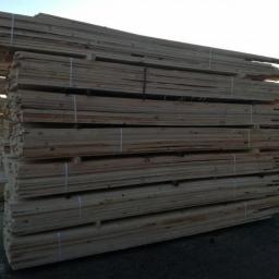 PPHU Ivbut - Stosy świeżo przetartego drewna budowlanego, zabezpieczone taśmami, składowane na zewnątrz, z zielonym trawnikiem i drzewami w tle.