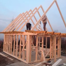 Konstrukcja szkieletowa budynku w trakcie montażu dachu, widoczni dwaj pracownicy na wysokości, surowe drewno, krajobraz w tle.