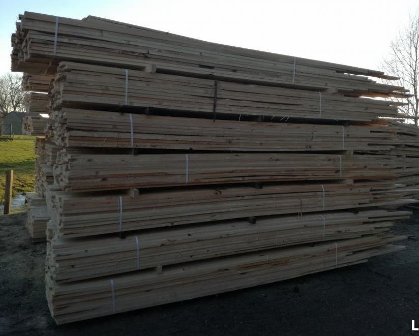 Stosy świeżo przetartego drewna budowlanego, zabezpieczone taśmami, składowane na zewnątrz, z zielonym trawnikiem i drzewami w tle.