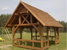 Drewniana altana ogrodowa z dwuspadowym dachem, widoczna konstrukcja więźby dachowej, na trawniku, w tle drzewa i huśtawka.