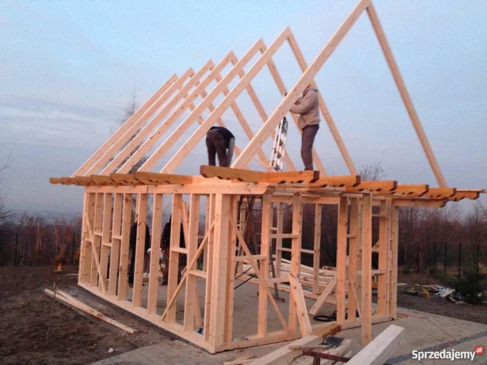 Konstrukcja szkieletowa budynku w trakcie montażu dachu, widoczni dwaj pracownicy na wysokości, surowe drewno, krajobraz w tle.