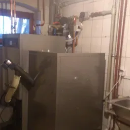 Szary kocioł grzewczy z rurami miedzianymi w kotłowni z białymi płytkami, widoczny fragment okna i umywalki.