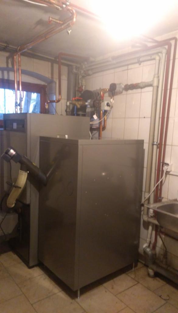 Szary kocioł grzewczy z rurami miedzianymi w kotłowni z białymi płytkami, widoczny fragment okna i umywalki.