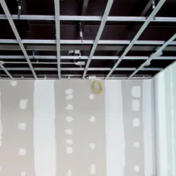 Montaż metalowej konstrukcji sufitu podwieszanego w pomieszczeniu z częściowo wykończonymi ścianami, widoczne przewody elektryczne.