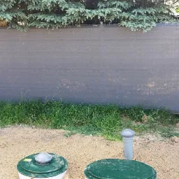 Dwa zbiorniki z zielonymi pokrywami i białą obudową, prawdopodobnie części instalacji oczyszczania ścieków, częściowo zasypane piaskiem, na tle żywopłotu i świerku