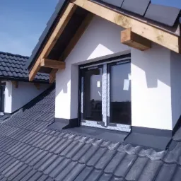 Nowy dach z ciemnej dachówki ceramicznej, lukarny z drewnianymi elementami konstrukcyjnymi i obróbkami blacharskimi, okna zabezpieczone folią ochronną.