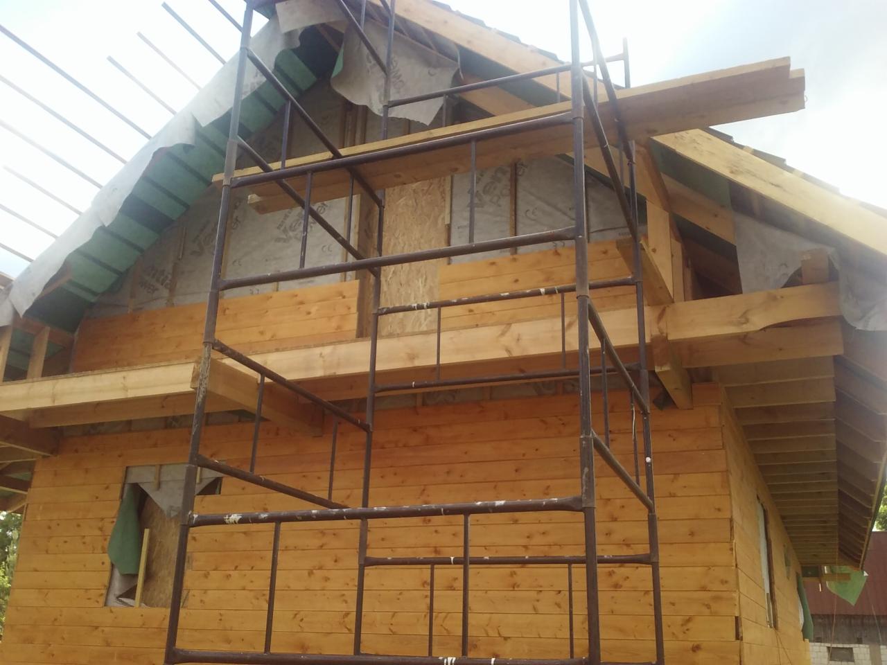 Budowa drewnianego domu w trakcie wznoszenia więźby dachowej, widoczne rusztowanie i elementy konstrukcyjne.