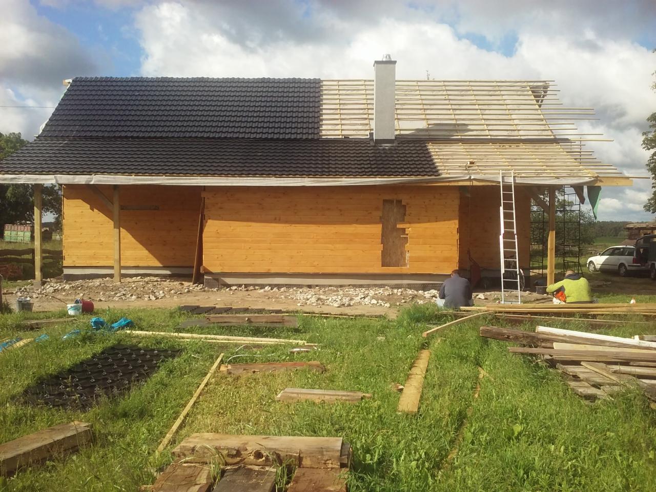Budowa domu z drewnianych bali: połowa dachu pokryta czarną dachówką, druga połowa w trakcie budowy więźby, widoczny komin, drabina i pracownicy.
