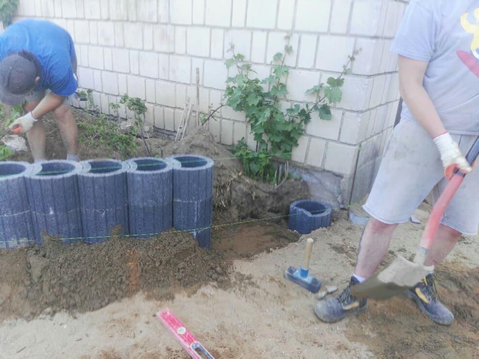 Prace ziemne przy budowie obrzeża rabaty kwiatowej z betonowych elementów, widoczni dwaj mężczyźni w trakcie pracy, narzędzia i poziomica na piasku.