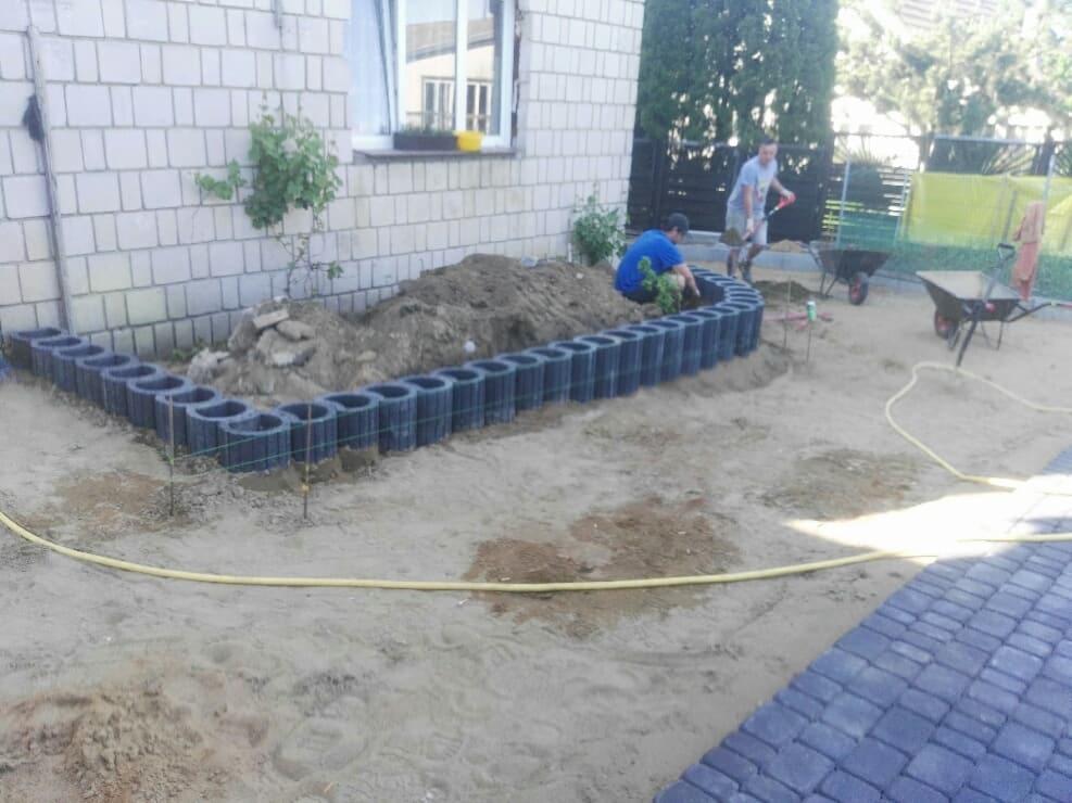 Prace ziemne przy obrzeżach rabaty kwiatowej wykonanej z betonowych elementów. W tle widoczna ściana budynku z oknem i pracownicy z taczkami.