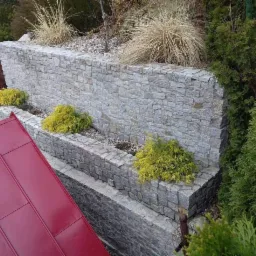 Trójstopniowa, kamienna ściana oporowa z żółtymi krzewami, widok z góry, fragment dachu w lewym dolnym rogu.