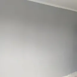 Świeżo pomalowana szara ściana z białą listwą przypodłogową, zabezpieczone drzwi wejściowe folią malarską, fragment drabiny malarskiej po prawej stronie.