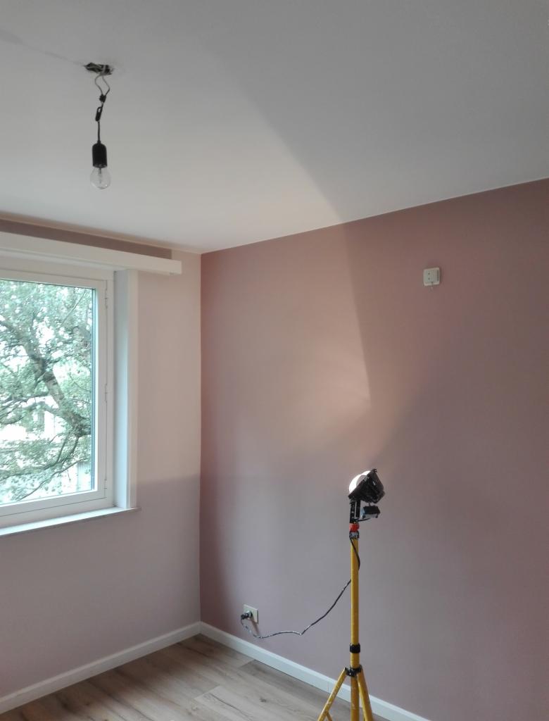 Pokój po malowaniu na dwa odcienie różu, widoczna lampa budowlana na trójnogu i surowa żarówka na kablu zwisająca z sufitu.