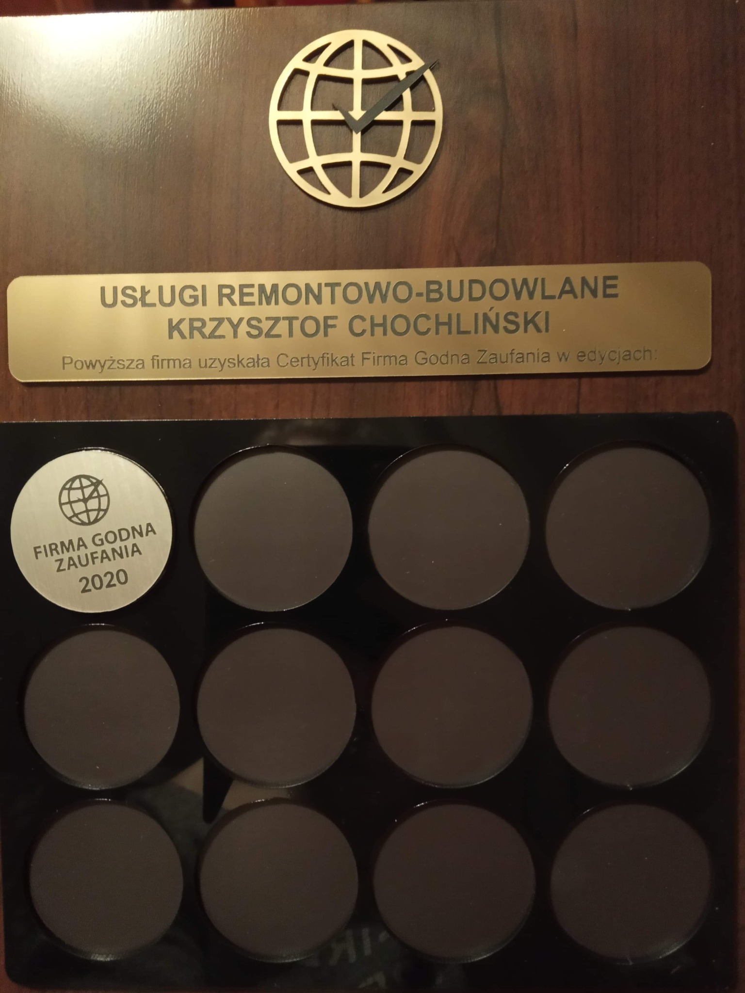 Tablica z logo firmy remontowo-budowlanej Krzysztof Chochliński oraz certyfikatem Firma Godna Zaufania 2020, umieszczona na drewnianej powierzchni z dekoracyjnym elementem w kształcie globu