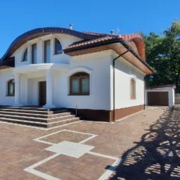 Elegancki, dwukondygnacyjny dom z białą elewacją, brązowym dachem i stolarką okienną, widoczny z zewnątrz w słoneczny dzień. Uwagę zwraca nietypowy, zaokrąglony kształt dachu nad wejściem, kolumny...